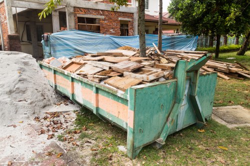 A large, heavy-duty green metal skip positioned on a grassy area near a residential construction site in Covent Garden, filled with irregularly stacked wooden planks, panels, and debris with a weathered, rough texture and natural wood tones. The skip is partially surrounded by fallen leaves and earth, with a small pile of construction material next to it. In the background, a partially built brick structure covered with blue plastic sheeting, and trees with green foliage are visible, indicating an outdoor environment consistent with the outskirts of the town. The scene is well-lit with natural daylight, conveying an active rubbish removal process typical for building or renovation waste management by Business Waste Removal Covent Garden, specialising in rubbish disposal services in the local area. The setting suggests a focus on construction debris disposal, with the skip set on the pavement beside a residential property and the surrounding environment reinforcing the context of waste collection and clearance services in central London's Covent Garden district.