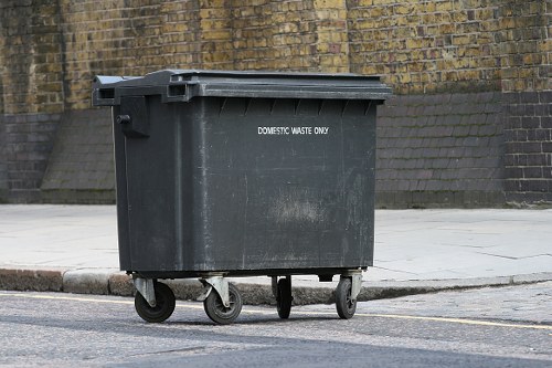 A large, dark grey plastic rubbish bin with a hinged lid marked 'DOMESTIC WASTE ONLY' is positioned on a paved sidewalk next to a curb stone in an urban street setting. The bin has four black wheels, with the front two slightly visible, supporting its rectangular shape. It is placed on a smooth, light grey pavement surface, adjacent to a curb that separates it from the tarmac road. The background features a brick wall with a weathered yellow-brown appearance, partially covered by a sloped concrete support at the base. The scene is lit by natural daylight, creating soft shadows around the bin, suggesting a typical day in a city area that could be in or near Covent Garden. The setting indicates a typical location for waste collection and rubbish removal services, with the bin awaiting collection or disposal, consistent with services provided by Business Waste Removal Covent Garden.