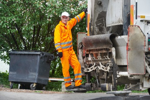 A waste collection operative dressed in bright orange high-visibility protective clothing and a white cap stands on a pavement near a large industrial rubbish compactor. The worker is smiling and positioned next to a partially open black wheeled waste bin, which is placed on the ground to the left of the individual. The compactor, made of metal with a dark, textured surface, is situated on a road or driveway, with a background of green leafy trees and bushes, suggesting a residential or urban environment. The scene is well-lit with natural daylight, displaying a clean, professional setting typical of rubbish removal operations in Covent Garden or nearby areas. Business Waste Removal Covent Garden provides such services, involving the collection and disposal of waste and rubbish in the local community.