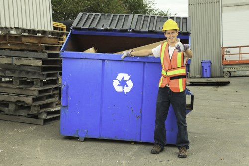 A male waste management worker wearing a yellow safety helmet, an orange high-visibility vest with reflective stripes, and dark trousers, standing outdoors on a paved surface. He is smiling and giving a thumbs-up with his right hand while holding a small piece of rubbish or a tool in his left hand. Next to him, there is a large blue skip with its lid open, revealing some waste inside. The skip features a white recycling symbol on its front and is positioned near a grey industrial building with metal walls. To the left of the skip, there are several stacked wooden pallets, and in the background, there are more industrial containers and equipment. The scene suggests an active rubbish collection or disposal operation, indicative of services offered by Business Waste Removal Covent Garden, serving the nearby local area, possibly in central London or the Covent Garden district.